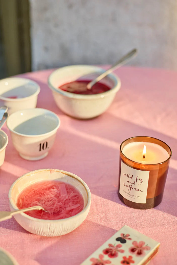 Candle with a label on a pink surface next to bowls of pink and white substances.