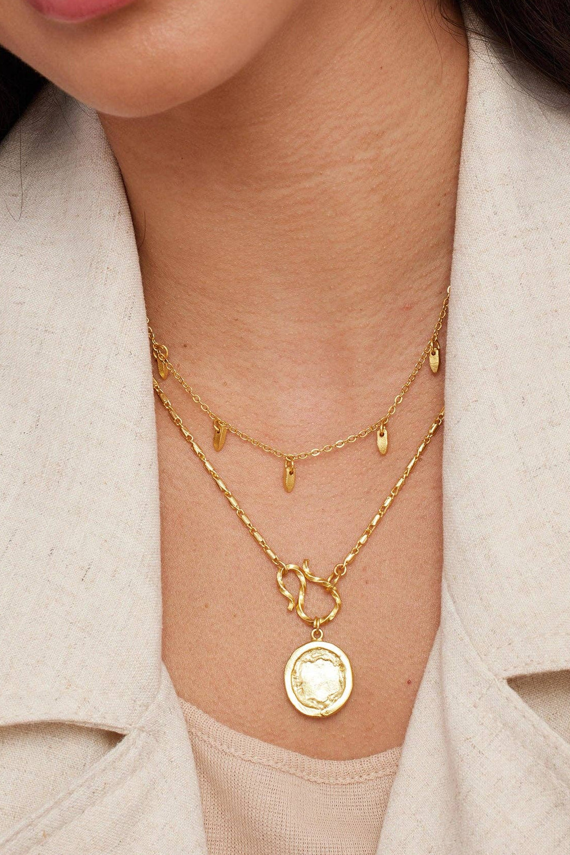 A close-up of a woman wearing layered gold necklaces and matching gold hoop earrings, styled with a light beige linen jacket against a solid pink background.
