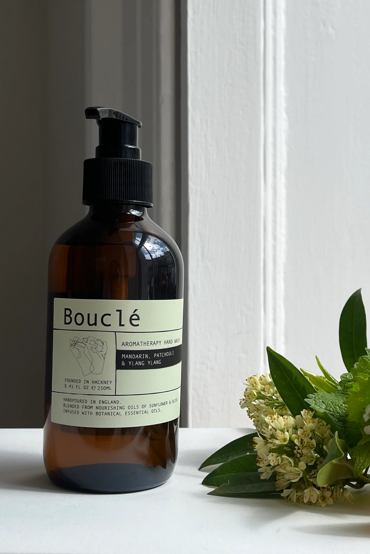 An amber glass pump bottle labeled Bouclé stands on a white surface beside green leaves, small white flowers, and an orange slice against a white paneled wall.
