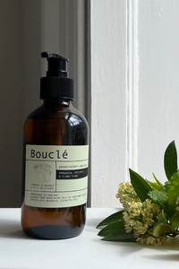 An amber glass pump bottle labeled Bouclé stands on a white surface beside green leaves, small white flowers, and an orange slice against a white paneled wall.