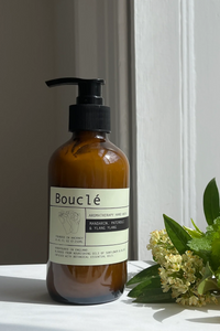 An amber glass pump bottle labeled Bouclé Aromatherapy Oil Series Manager & Plant Oil stands on a white surface beside green leaves, small white flowers, and orange slices in natural light.