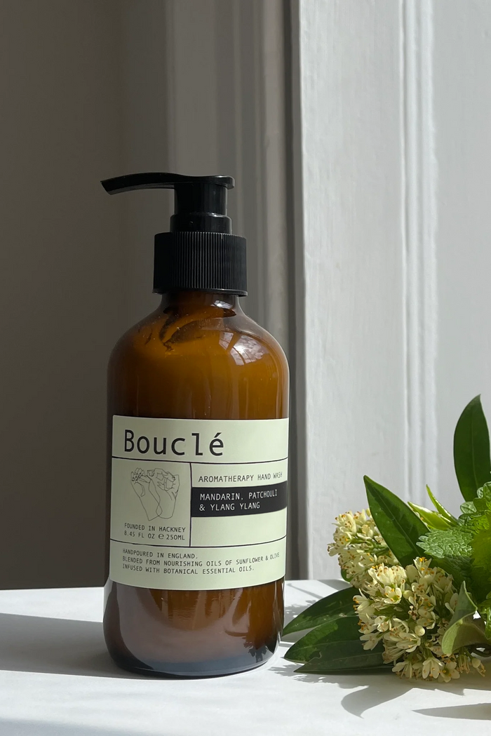 An amber glass pump bottle labeled Bouclé Aromatherapy Oil Series Manager & Plant Oil stands on a white surface beside green leaves, small white flowers, and orange slices in natural light.