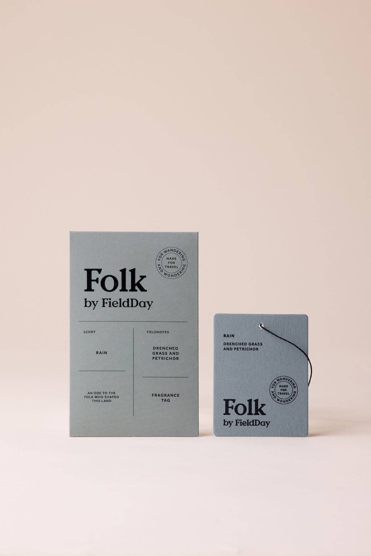 A minimalist fragrance product set on a warm beige background, featuring a tall rectangular box and a smaller square packet both printed with the brand name 'Folk by FieldDay' in black text on a muted blue-gray surface.