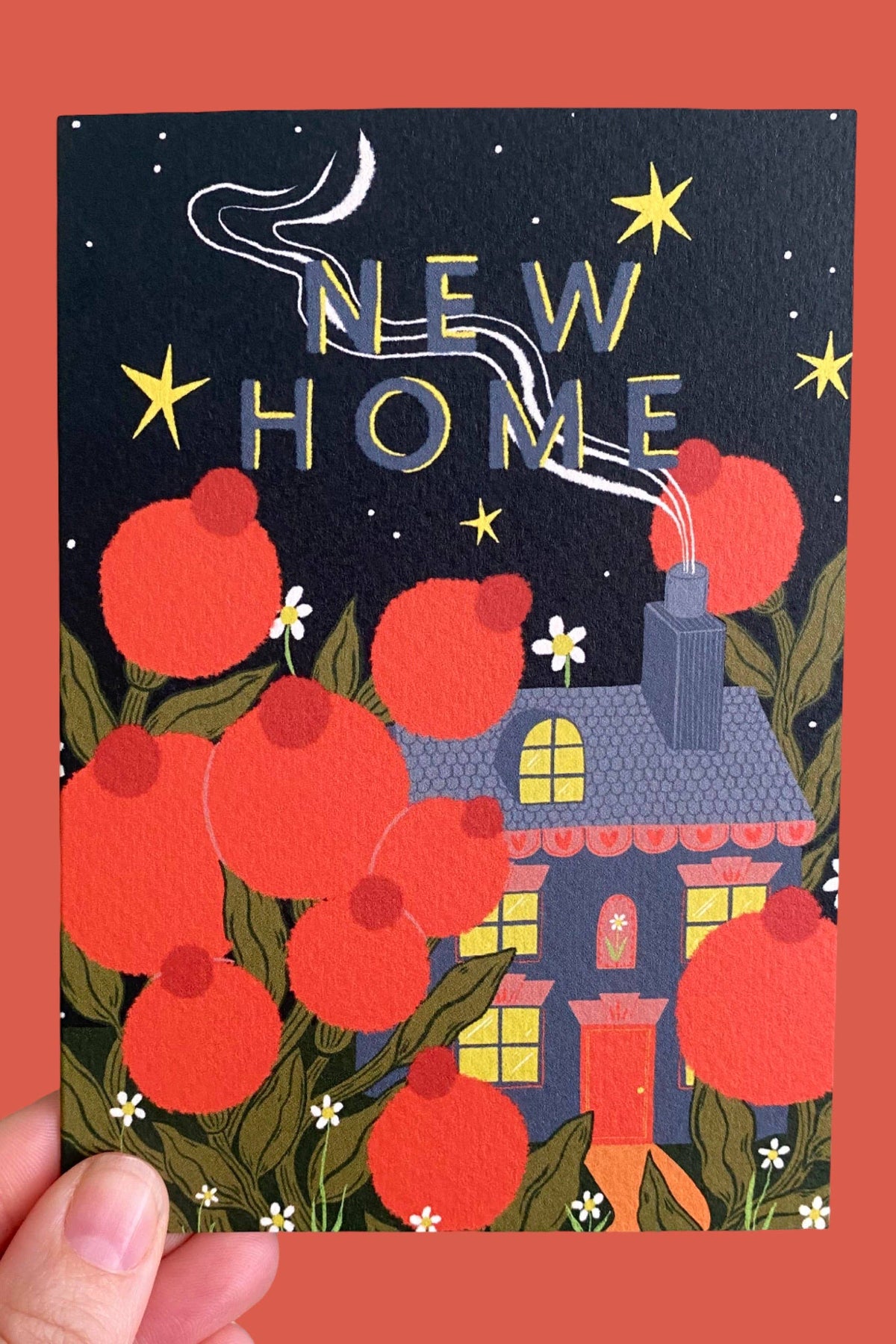 A hand holds a square greeting card featuring a whimsical illustration of a house, large red flowers, green leaves, and the words 'NEW HOME' in gold foil against a dark night sky. The card is shown against a solid coral-red background.