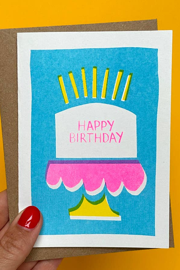 Hand holding a birthday card with a cake illustration on a yellow background