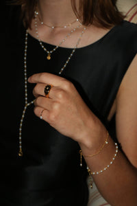 A close-up lifestyle shot of a person wearing layered gold and pearl necklaces, a beaded bracelet, and a black ring with a central white stone.