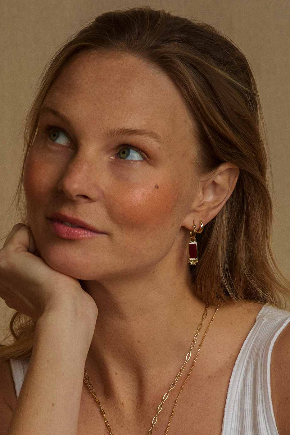 A close-up portrait of a woman with blonde hair, wearing a gold huggie hoop earring with a dark red rectangular drop pendant, layered with a delicate gold chain necklace, against a warm beige background.