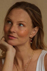A close-up portrait of a woman with blonde hair, wearing a gold huggie hoop earring with a dark red rectangular drop pendant, layered with a delicate gold chain necklace, against a warm beige background.