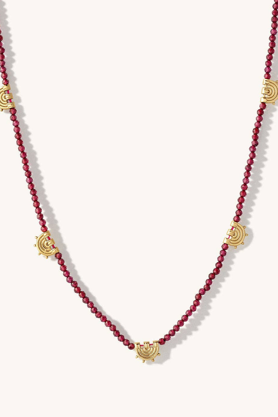 A beaded necklace featuring deep red round beads and five gold-toned sunburst charms spaced along the strand, displayed on a light cream background.