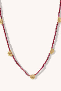 A beaded necklace featuring deep red round beads and five gold-toned sunburst charms spaced along the strand, displayed on a light cream background.