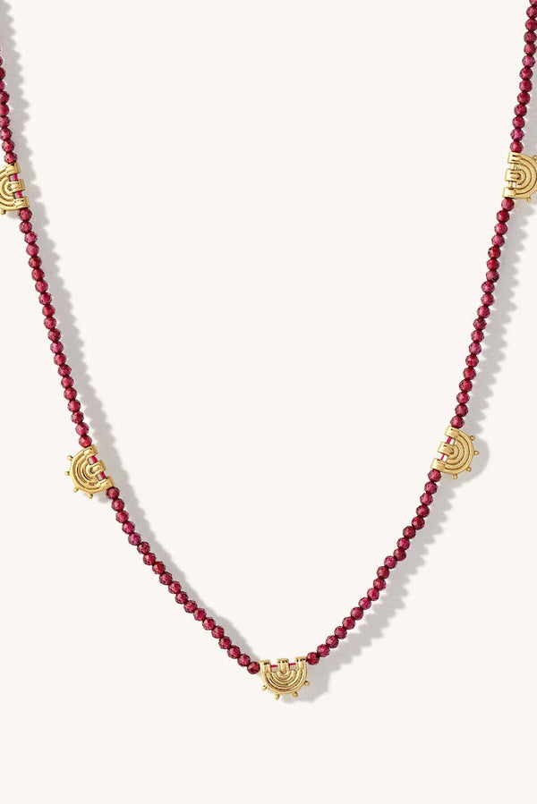 A beaded necklace featuring deep red round beads and five gold-toned sunburst charms spaced along the strand, displayed on a light cream background.