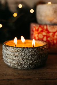 A close-up of a lit candle in a textured, speckled gray ceramic holder with three wicks, set on a rustic wooden surface with warm bokeh lights in the background.