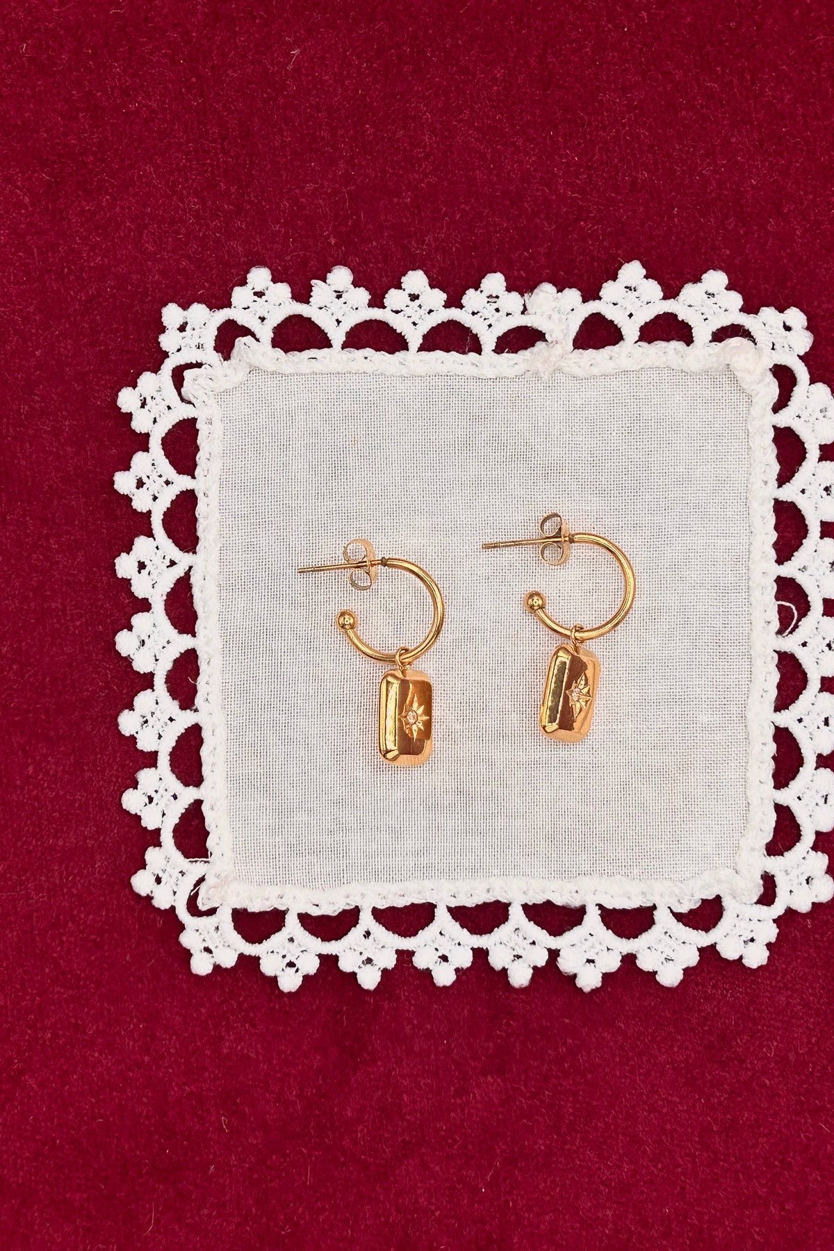 A pair of small gold hoop earrings with dangling polished gold charms, displayed on a white fabric square with a white lace border against a deep red background.