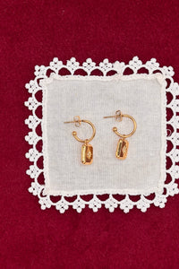A pair of small gold hoop earrings with dangling polished gold charms, displayed on a white fabric square with a white lace border against a deep red background.