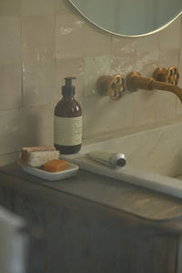 Bottle of soap on a soap dish next to a sink with a gold faucet.