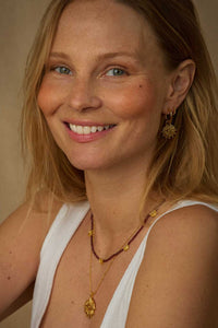 A smiling blonde woman wearing gold jewelry, including a gold pendant necklace, a beaded necklace, and a gold sunburst earring, against a warm beige background.