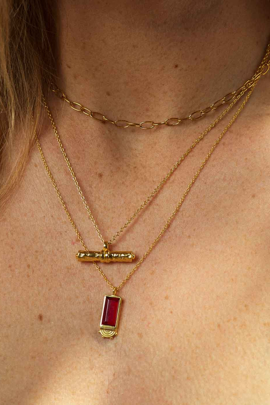 A close-up of a person's neck and chest wearing layered gold necklaces, including a curb chain, a delicate chain with a small gold bar pendant, and a longer chain with a rectangular red gemstone pendant.