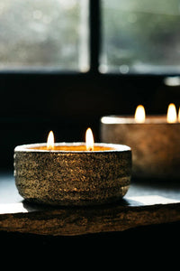 Two textured ceramic candle holders with lit candles, placed on a dark windowsill with a blurred window and outdoor view in the background.