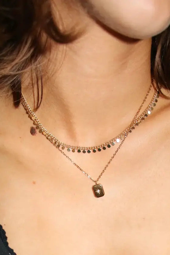 A close-up of a model wearing a layered gold necklace with a small pendant, paired with a black and white polka dot top.