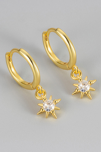 A pair of gold huggie hoop earrings with dangling starburst charms set with clear round gemstones, shown on a neutral light gray background.