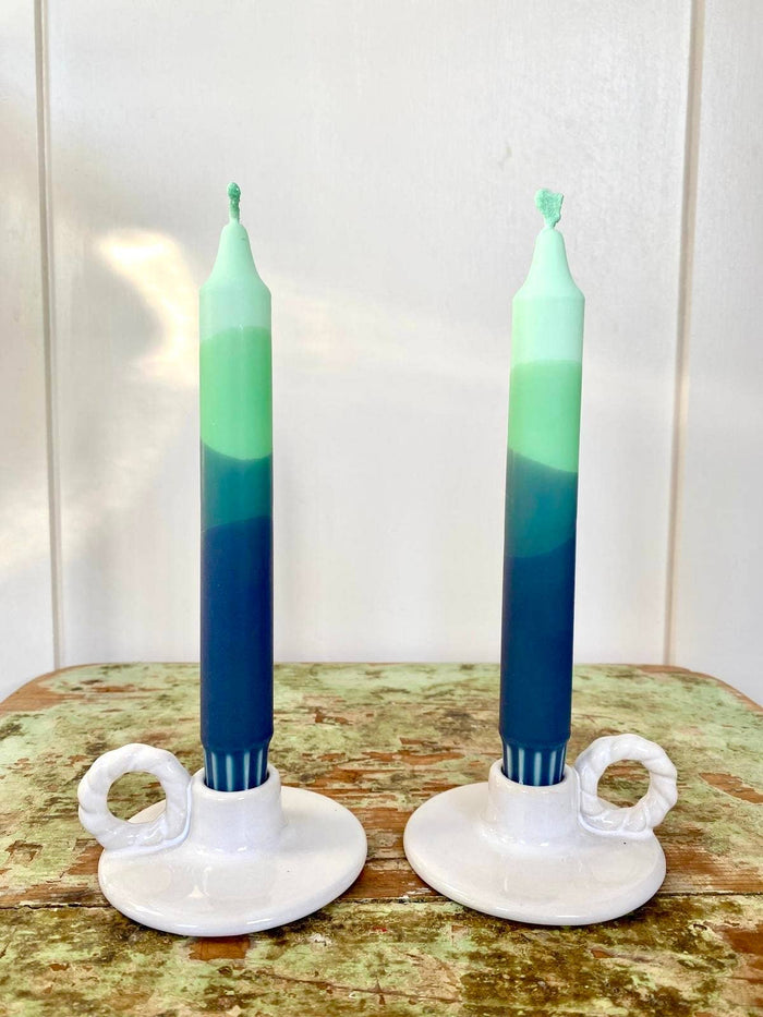 Two green and blue gradient candles in white holders on a wooden surface with a white background.