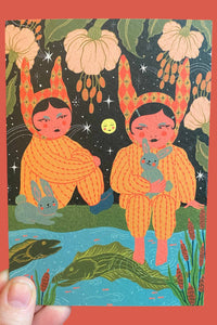A hand holds a square art print featuring a whimsical illustration of two children in orange bunny suits sitting by a pond under a starry night sky with pumpkins and flowers. The print is set against a solid coral background.