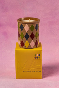 Candle in a patterned container on top of a yellow box with a pink background