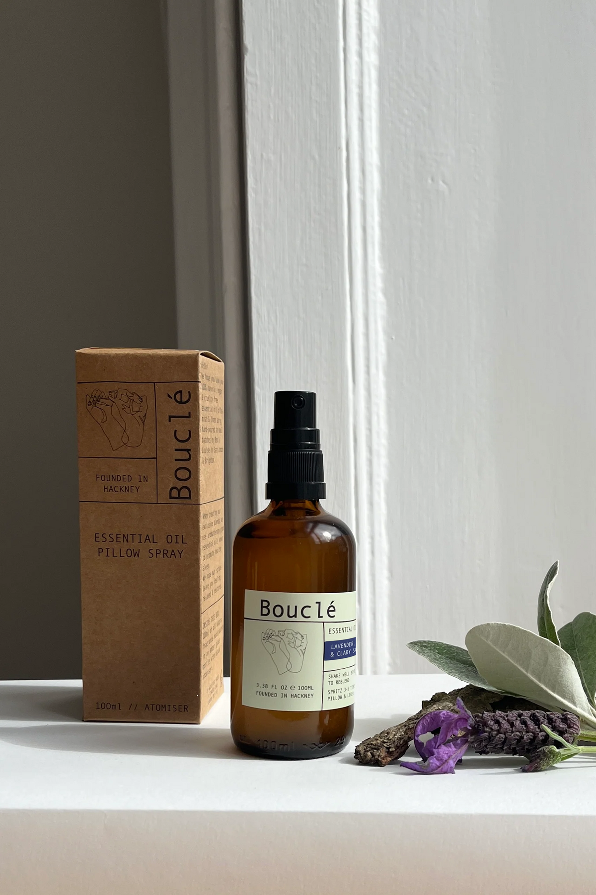 Bouclé Essential Oil Pillow Spray bottle in an amber glass spray bottle with a black cap, standing next to its brown cardboard box on a white windowsill with lavender and green leaves in the foreground.