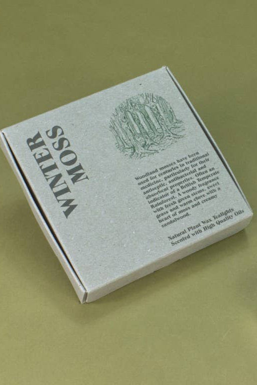 A silver-gray box labeled 'Winter Moss' with three small white tea light candles arranged beside it on a solid olive-green background.