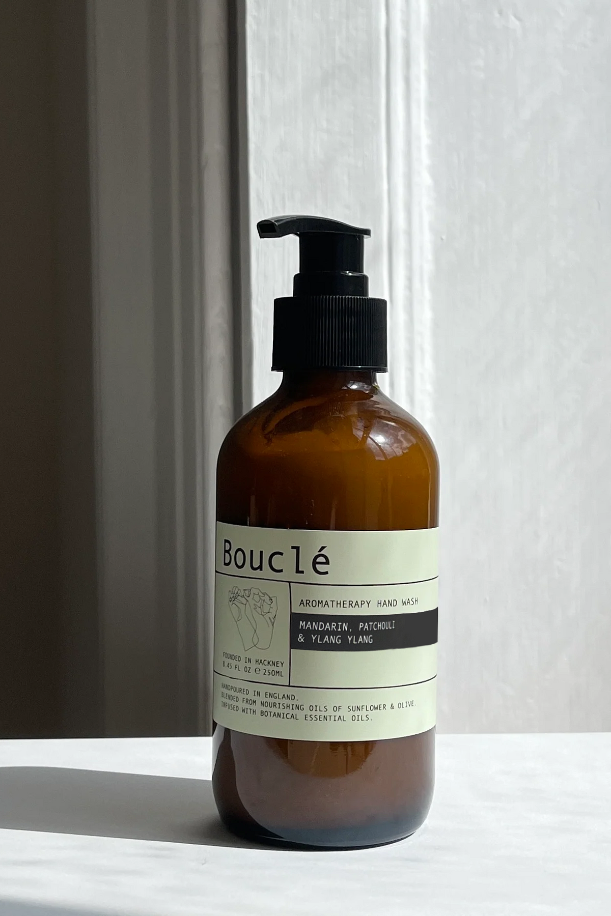 An amber glass pump bottle with a black dispenser and a cream label reading Bouclé, set on a white surface against a white paneled wall with soft natural light.