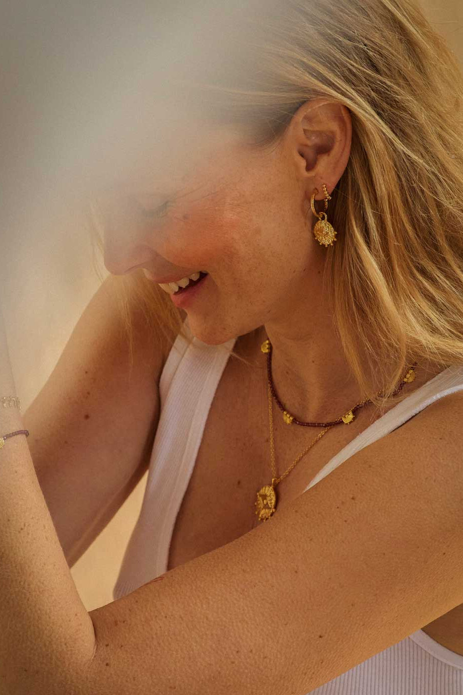 A close-up lifestyle shot of a woman wearing gold jewelry, including a gold dangle earring, layered gold necklaces, and a gold bracelet, paired with a white tank top.