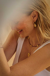 A close-up lifestyle shot of a woman wearing gold jewelry, including a gold dangle earring, layered gold necklaces, and a gold bracelet, paired with a white tank top.