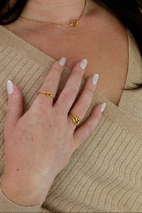 A close-up lifestyle shot of a woman wearing delicate gold rings on her fingers, a thin gold necklace, and a beige ribbed knit top.