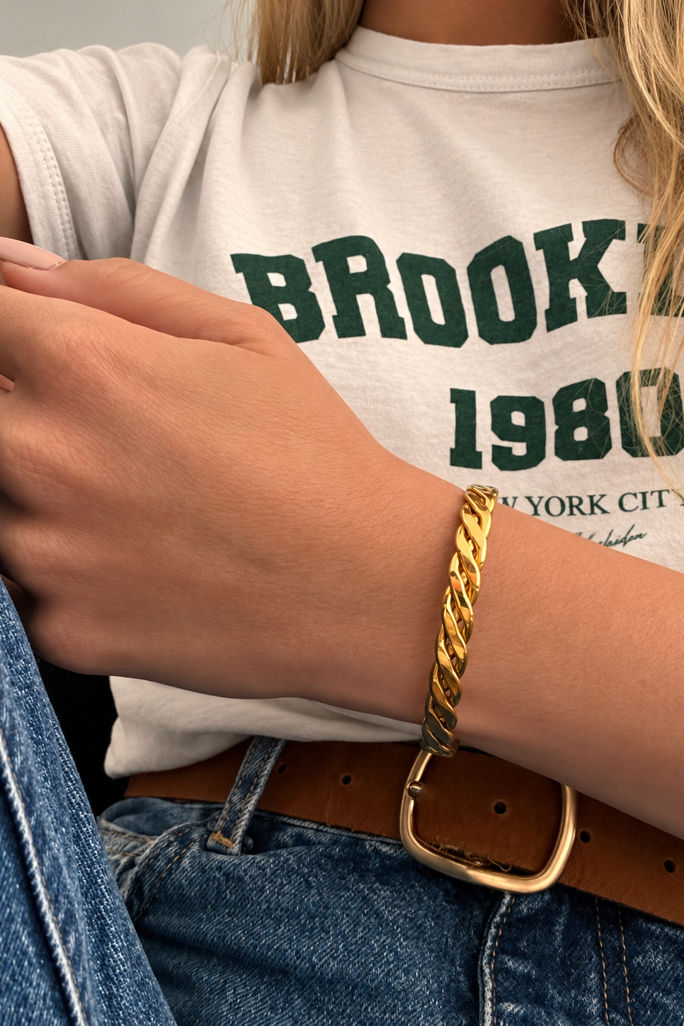 A close-up of a person wearing a gold chain-link bangle bracelet on their wrist, styled with a white graphic t-shirt and blue jeans.