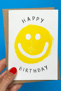 Hand holding a birthday card with a yellow smiley face and 'Happy Birthday' text against a blue background.
