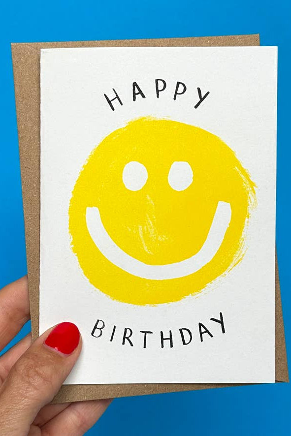 Hand holding a birthday card with a yellow smiley face and 'Happy Birthday' text against a blue background.