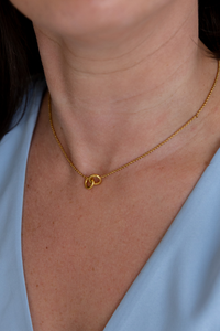 A close-up of a model's neck and chest wearing a delicate gold chain necklace with a small interlocking rings pendant, paired with a light blue top.