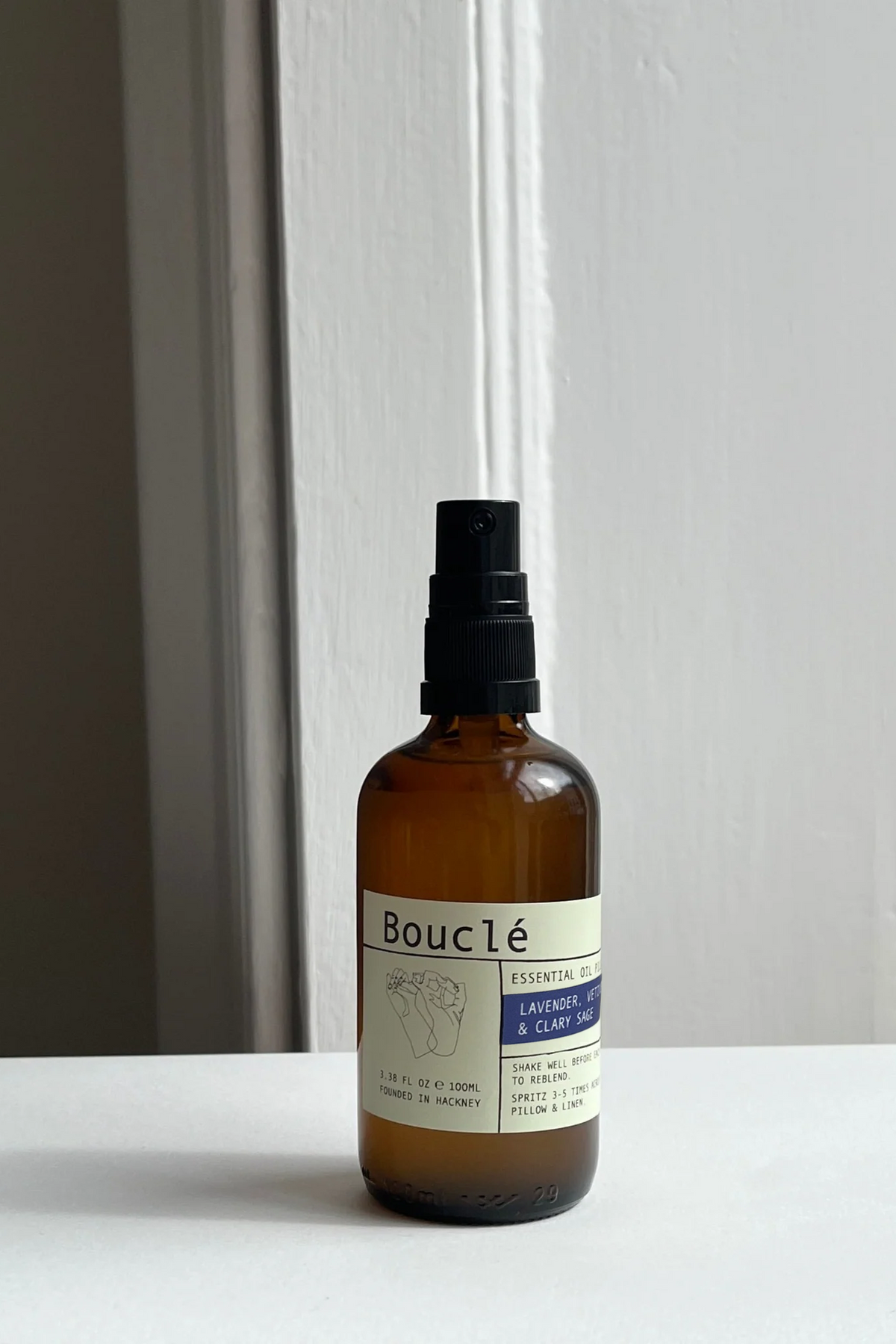 A bottle of Bouclé Lavande & Clâme essential oil spray, made from an amber glass bottle with a black spray nozzle and a cream-colored label, set on a white surface against a white paneled wall.