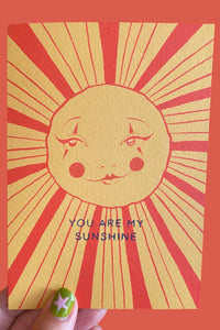 A hand holds a greeting card featuring a stylized sun face with radiating red lines on a yellow background and the text 'YOU ARE MY SUNSHINE'. The card is shown against a solid coral-red background.