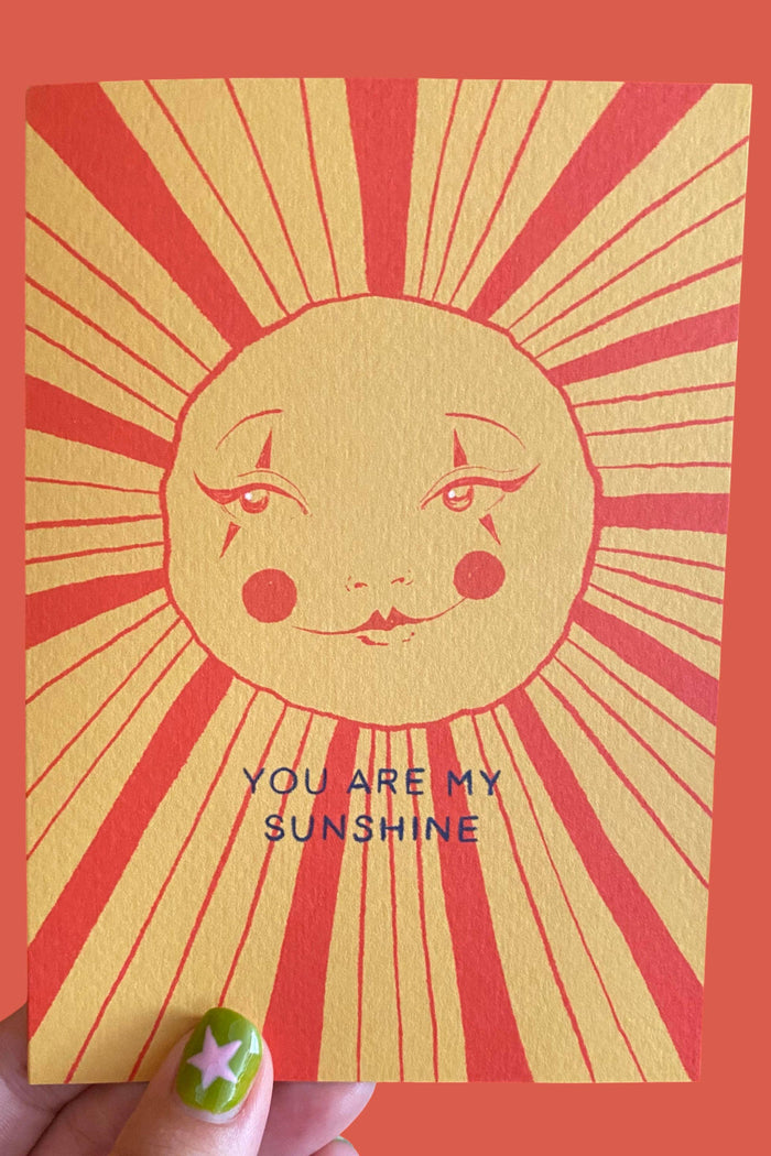 A hand holds a greeting card featuring a stylized sun face with radiating red lines on a yellow background and the text 'YOU ARE MY SUNSHINE'. The card is shown against a solid coral-red background.