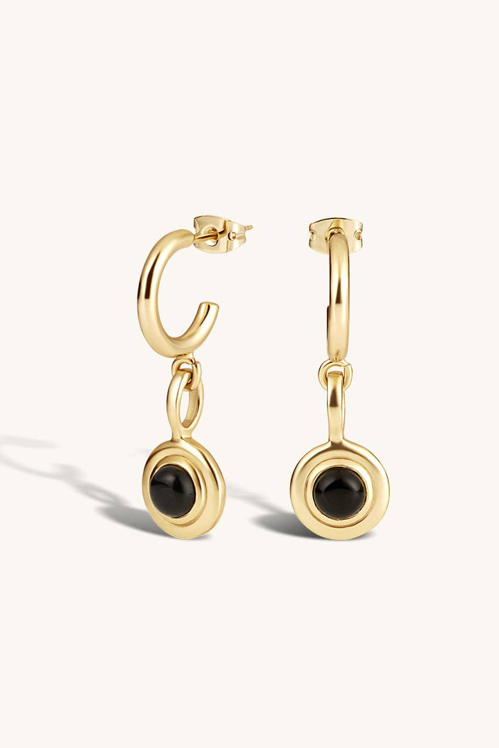 A pair of gold huggie hoop earrings with a dangling circular disc featuring a black onyx stone, shown on a clean off-white background.