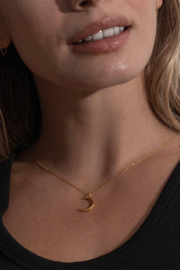 A close-up of a woman wearing a delicate gold crescent moon pendant necklace and a small gold hoop earring, paired with a black ribbed tank top.