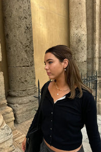 A woman wearing a black cropped button-up cardigan over a white top, standing in front of large stone columns.
