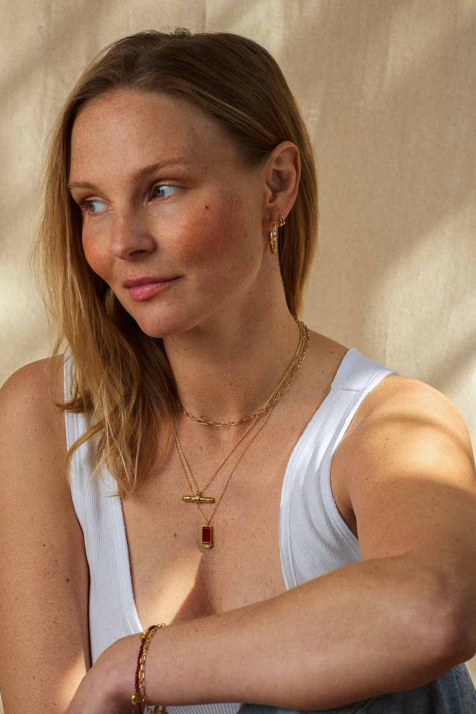 A woman wearing layered gold necklaces, one with a small red rectangular pendant, and gold bracelets, posing in a white tank top against a warm beige textured backdrop.