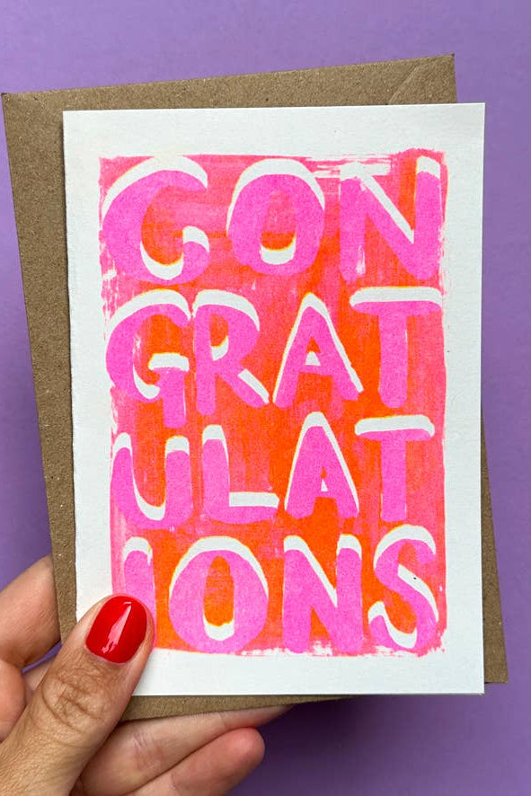 Hand holding a card with 'CONGRATULATIONS' in pink and orange on a purple background