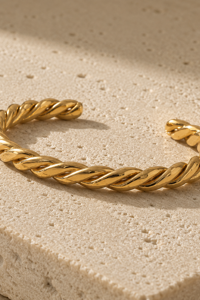A twisted rope-style cuff bracelet in polished yellow gold, resting on a textured beige stone surface with soft shadows.