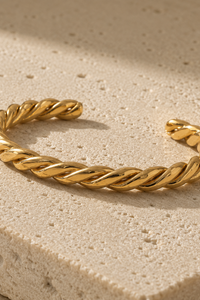 A twisted rope-style cuff bracelet in polished yellow gold, resting on a textured beige stone surface with soft shadows.
