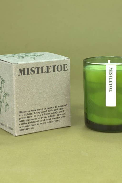A green glass jar candle next to its light gray box, both labeled 'MISTLETOE', on a solid olive green background.