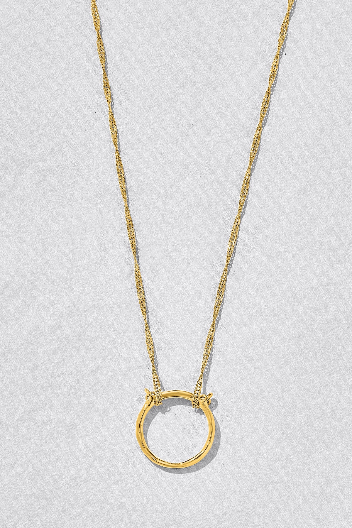 A delicate gold chain necklace featuring a large, textured open circle pendant, displayed against a light gray textured background.