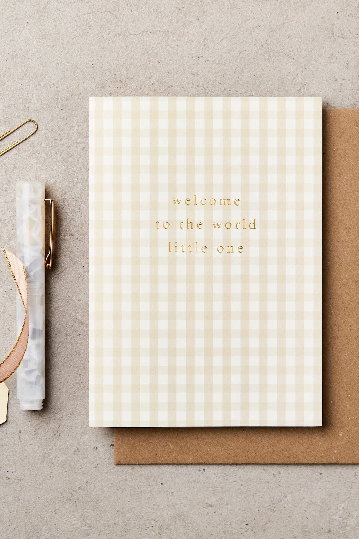 Greeting card with 'welcome to the world little one' text on a checkered pattern, with a brown envelope and stationery items on a beige background.